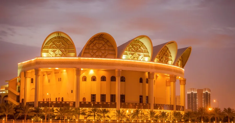 Das Bahrain National Theatre in der Abenddämmerung golden beleuchtet am Wasser, Palmen am Ufer